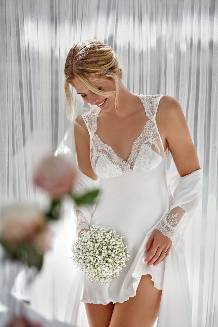 A woman wearing a white bridal chemise with lace details and long sleeves, holding a bouquet of white flowers.